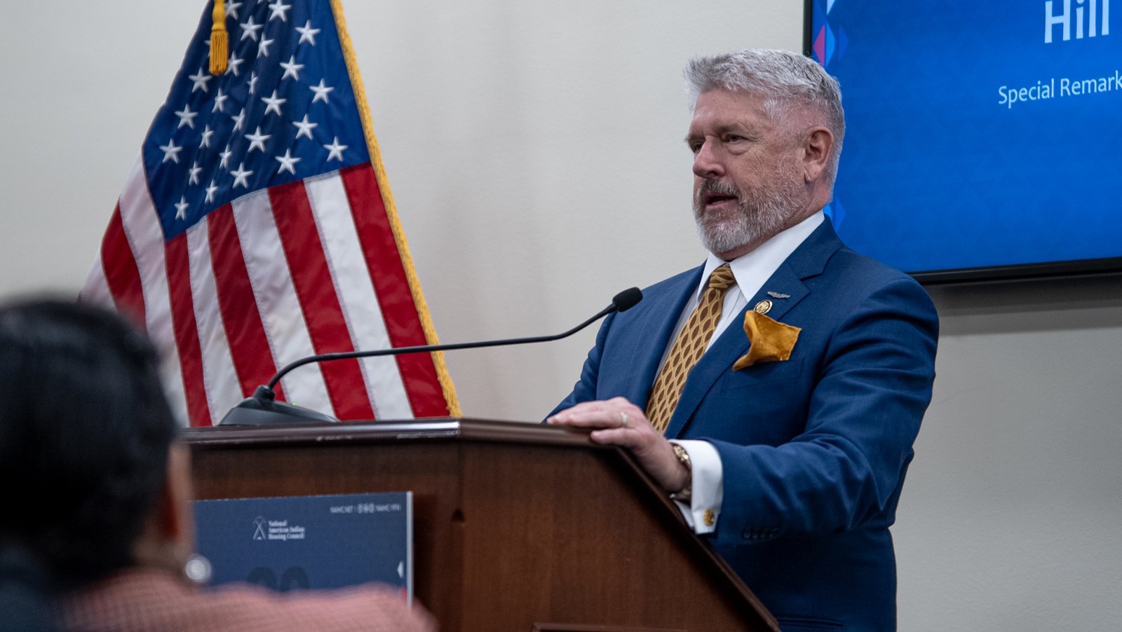 Rep. Troy Downing speaking at a podium beside the American flag during NAIHC Hill Meetings